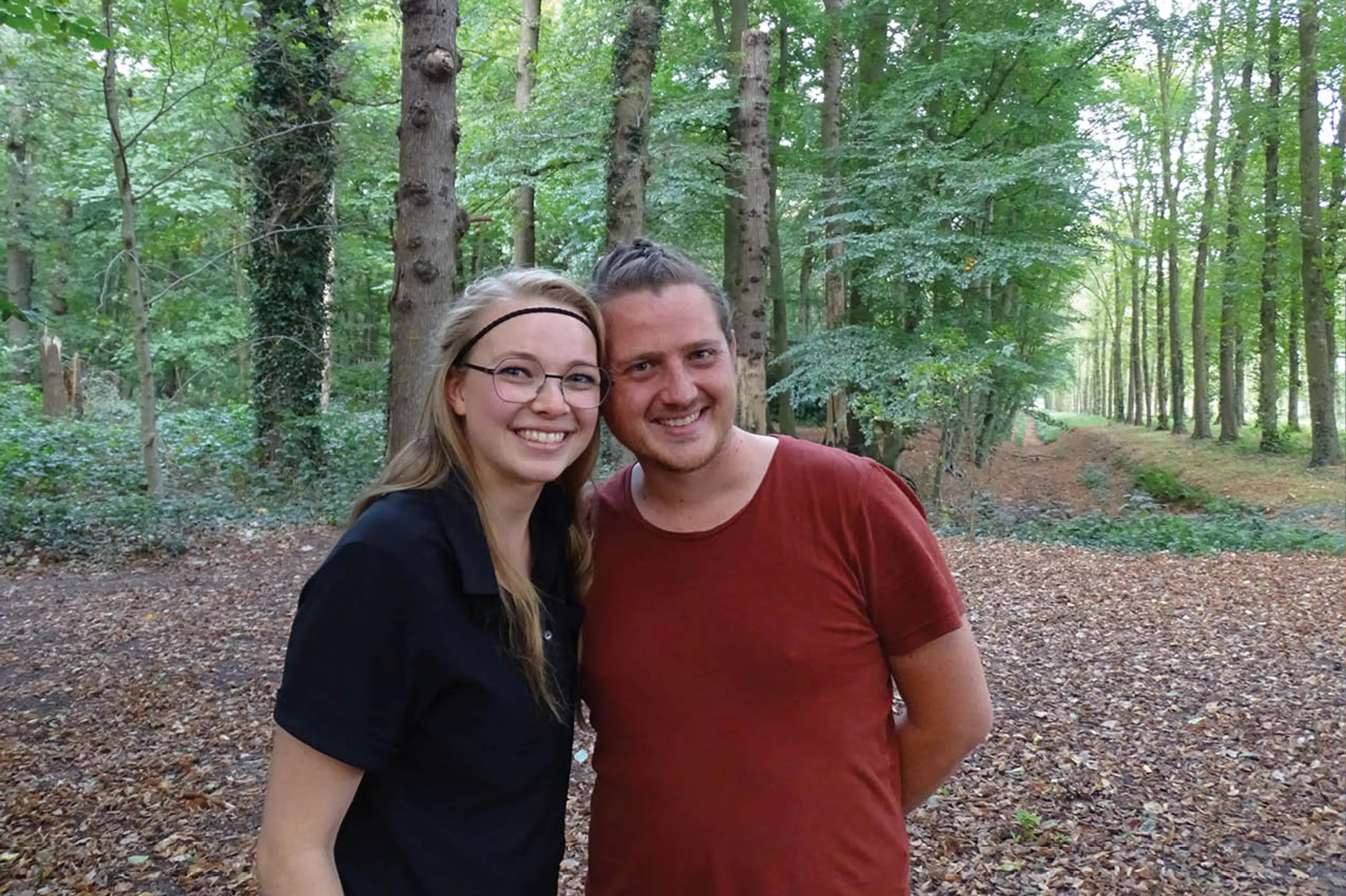 Saskia en Hilbert staan in een bos. Ze lachen in de camera. Hilbert draagt een donderrood shirt, en Saskia een zwart shirt